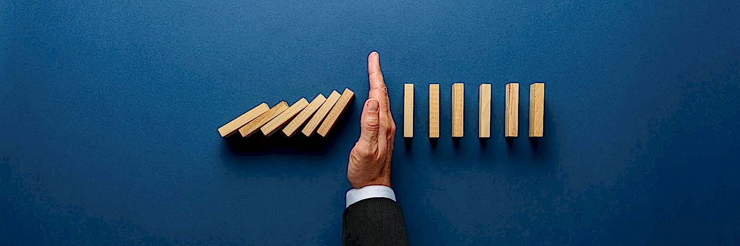 Wide view image of businessman hand stopping collapsing dominos in a conceptual image. Top view over navy blue background.